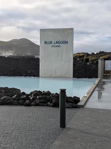 The Blue Lagoon entrance sign beside milky blue water and black lava rock